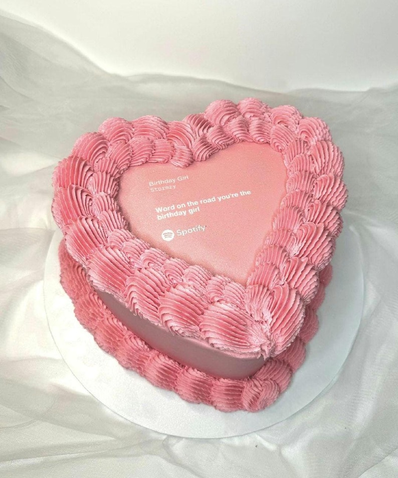Pode incluir: Um bolo rosa em forma de cora&ccedil;&atilde;o com uma borda de glac&ecirc; texturizada. O centro do bolo tem uma superf&iacute;cie lisa com o texto "Birthday Girl Stories" e "Word on the road you're the birthday girl" com o logotipo do Spotify.
