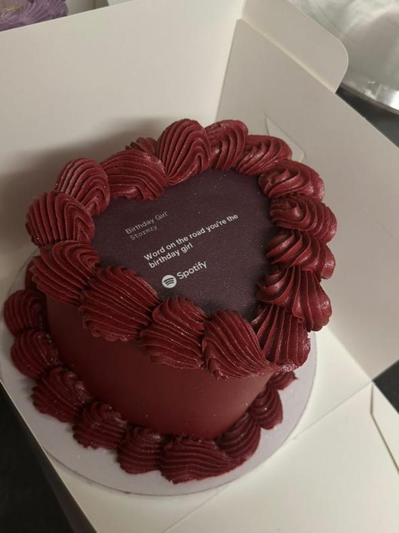 Pode incluir: Um bolo redondo, bord&ocirc;, com redemoinhos de glac&ecirc; vermelho escuro nas bordas. O centro do bolo tem uma superf&iacute;cie roxa escura com texto branco: "Birthday Girl Stoxmzy" e "Word on the road you're the birthday girl" com o logotipo do Spotify.