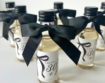 May include: Miniature glass bottles filled with a light-colored liquid, each adorned with a black bow. The bottles have black caps and white labels that read "Cheers to 30+". The bottles are arranged on a white surface.