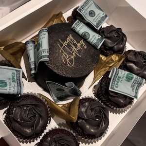 Può includere: Una scatola di cupcake al cioccolato e una piccola torta decorata per un compleanno. I cupcake hanno una glassa scura a forma di rosa. La torta è nera con testo dorato che dice "Happy Birthday". Banconote da $100 arrotolate sono poste intorno alla torta e ai cupcake.