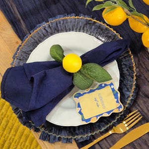 Puede incluir: Una servilleta de lino azul marino está doblada y adornada con un limón amarillo y hojas verdes, colocada sobre un plato blanco. El plato se asienta sobre un cargador transparente con borde dorado. Un tenedor y un cuchillo dorados están a la derecha.
