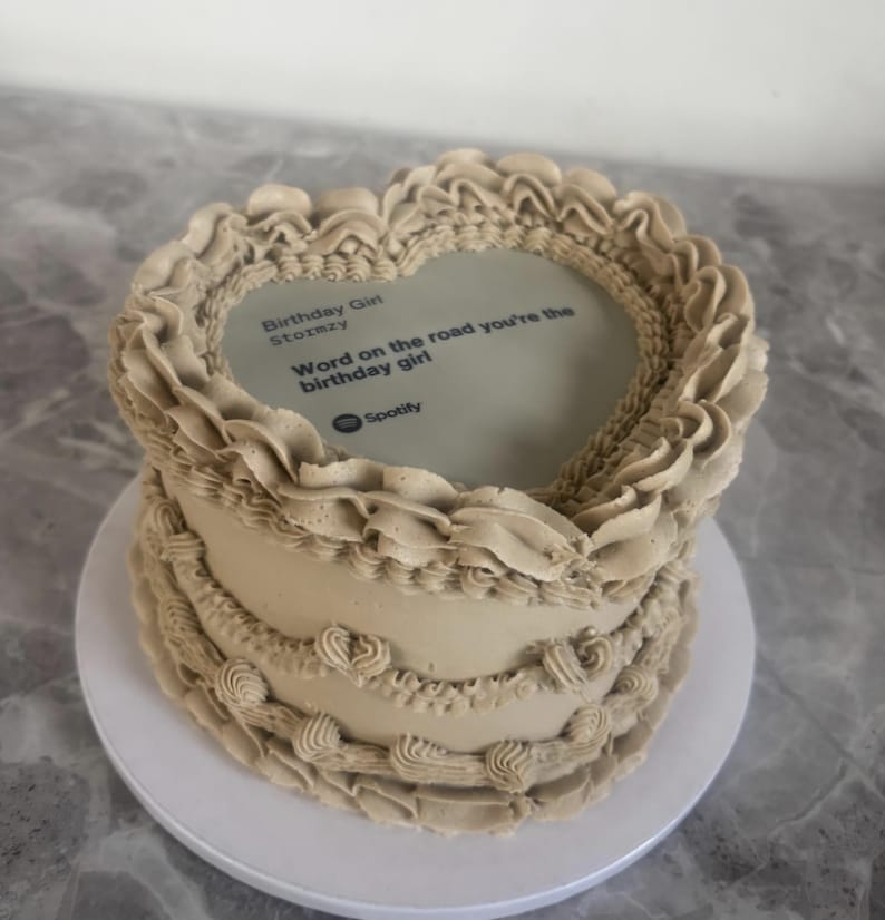 Pode incluir: Bolo redondo, com cobertura bege, com decora&ccedil;&otilde;es de creme nas bordas. A parte superior do bolo apresenta uma imagem comest&iacute;vel em forma de cora&ccedil;&atilde;o com o texto "Birthday Girl Stormzy Word on the road you're the birthday girl" e o logotipo do Spotify.