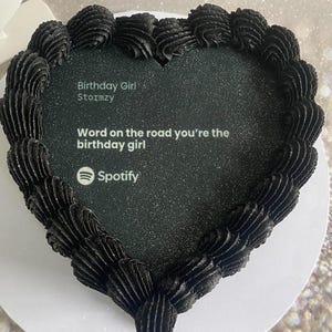 Pode incluir: Um bolo em forma de cora&ccedil;&atilde;o com cobertura preta e uma superf&iacute;cie preta brilhante. O bolo tem o texto "Birthday Girl Stormzy" e "Word on the road you're the birthday girl" com o logotipo do Spotify.