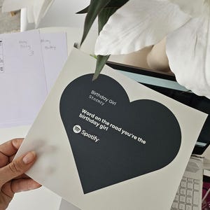 Pode incluir: Um cart&atilde;o branco com um design em forma de cora&ccedil;&atilde;o preto. O cart&atilde;o apresenta o texto "Birthday Girl Stormzy" e "Word on the road you're the birthday girl" com o logotipo do Spotify. O cart&atilde;o &eacute; segurado por uma m&atilde;o.