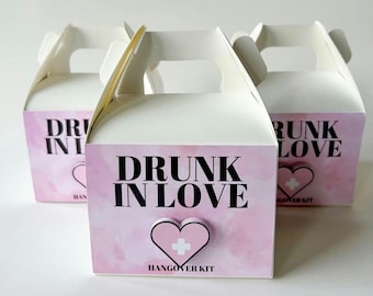 May include: Three white gift boxes with handles, each featuring a pink front panel with the words "DRUNK IN LOVE" in black. A pink heart with a white cross is below the text, with "HANGOVER KIT" underneath.