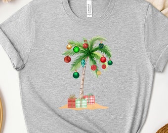 May include: A gray t-shirt with a Christmas palm tree design. The palm tree is decorated with red, green, and gold ornaments and has presents at the base.
