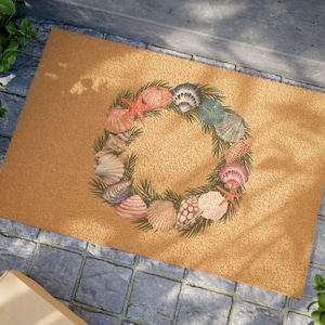 May include: A brown coir doormat with a wreath of seashells and pine branches. The wreath is made up of various seashells in shades of pink, white, and brown, as well as a starfish and a sand dollar.