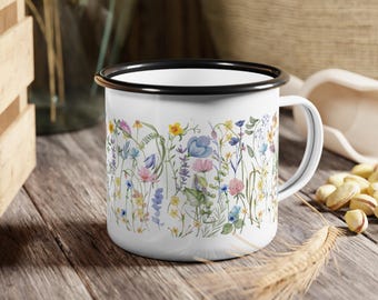 May include: White enamel mug with a black rim and handle, decorated with a colorful floral design. The wildflowers include yellow, blue, pink, and purple blooms. The mug is sitting on a wooden surface.