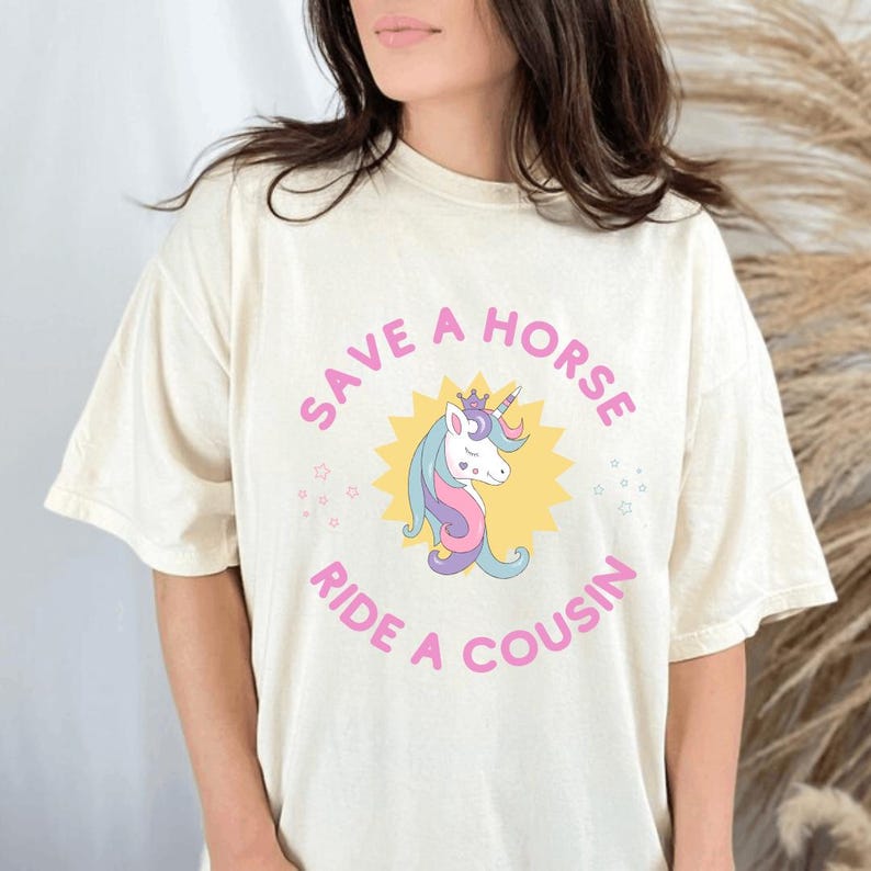 May include: Off-white t-shirt featuring the slogan "SAVE A HORSE RIDE A COUSIN" in pink, with a cartoon unicorn graphic. The unicorn has a crown, a pink and purple mane, and a yellow starburst background.
