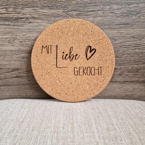 Könnte beinhalten: Ein runder Korkuntersetzer mit dem Text "MIT LIEBE GEKOCHT" und einem Herzsymbol.