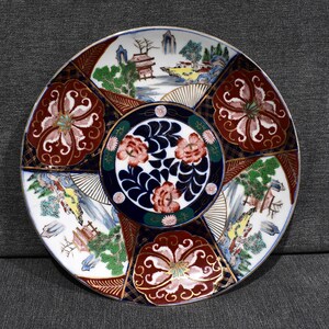 May include: A round ceramic plate with a white background and intricate designs in red, blue, gold, and green. The plate features floral patterns, stylized trees, and a central motif of a pagoda-like structure.