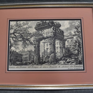 May include: A black and white engraving of a Roman temple ruin. The engraving is framed in a gold and silver frame. The text below the image reads "Avanzo del Pronao del Tempio di Giove Tonante. A. Avanzi del Tabulario. Piranesi Architetto."
