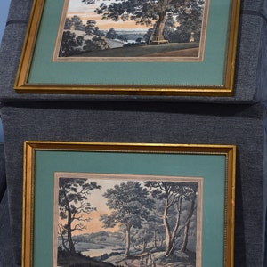 May include: Two framed landscape paintings with gold frames. The top painting depicts a tree with a bench in the foreground and a river in the background. The bottom painting depicts a river with trees and a path leading to a group of people in the distance.