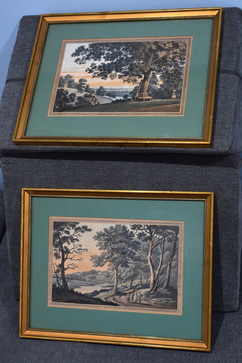 May include: Two framed landscape paintings with gold frames. The top painting depicts a tree with a bench in the foreground and a river in the background. The bottom painting depicts a river with trees and a path leading to a group of people in the distance.