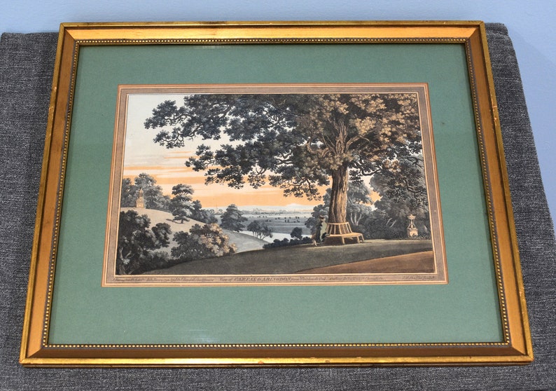 May include: A framed print of a landscape scene with a large tree in the foreground, a winding path, and a distant town. The print is in shades of brown, green, and blue, and has a gold frame.