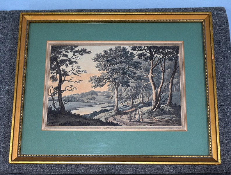May include: A framed print of a landscape scene with trees, a river, and figures in the distance. The print is in a gold frame with a green mat.