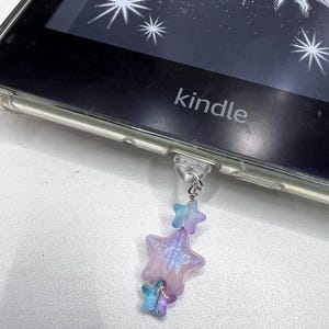 Könnte beinhalten: Ein violetter, blauer und schillernder sternförmiger Anhänger hängt an einem Kindle E-Reader. Der Anhänger ist mit einem durchsichtigen Plastikring an der Unterseite des E-Readers befestigt.