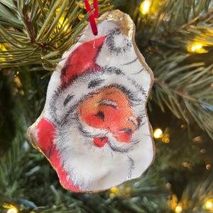 May include: A Christmas ornament featuring Santa Claus's face, painted on a white, shell-shaped surface. The ornament has a red hat and a gold-painted edge, with a red ribbon for hanging. The ornament is hanging on a Christmas tree.
