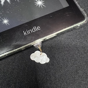 May include: A white cloud charm hanging from a Kindle e-reader. The e-reader is black with the word "kindle" printed on it. The cloud charm is attached with a gold-colored chain.