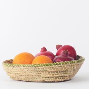 May include: A woven basket with a green trim holds oranges and pomegranates. The oranges are bright orange, and the pomegranates are a deep red. The basket is made of natural fibers.