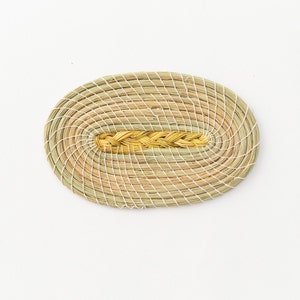 May include: A woven oval-shaped coaster made of natural materials. The coaster has a braided design in the center and is a light brown color.