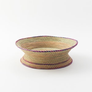 May include: A woven basket with a wide, round base and a flared rim. The basket is made of natural straw and has a pink and blue trim.