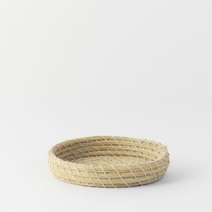 May include: A round, woven basket made of natural-colored straw. The basket has a shallow depth and a tightly woven, diamond-patterned design. The rim is slightly raised, and the basket sits on a white background.