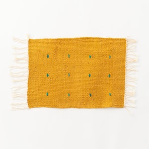 May include: Mustard yellow woven rug with teal diamond-shaped accents. The rug has a rectangular shape with a fringe on two sides. The texture of the rug appears to be coarse and handmade, suitable for home decor.