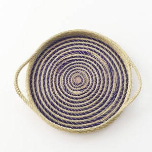 May include: A round woven tray with a purple and natural straw design. The tray has a spiral pattern and two handles.