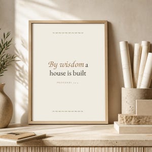 以下が含まれることがあります： 明るいクリーム色の背景に、ダークブラウンのセリフ体で「By wisdom a house is built」および「Proverbs 24:3」と記された長方形の額入りウォールアートプリント。上下に2本の細い水平な装飾線が施されています。