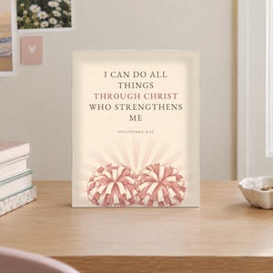 May include: A rectangular sign with a cream background featuring the text I CAN DO ALL THINGS THROUGH CHRIST WHO STRENGTHENS ME PHILIPPIANS 4:13 in dark brown lettering, with two pink and white pom-poms illustrated at the bottom center.