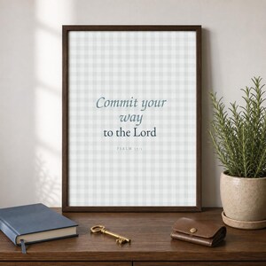 May include: A framed wall art print featuring a light grey and white gingham pattern background with the dark blue text Commit your way to the Lord and Psalm 37:5 printed in the centre. The frame is a dark brown wood with a simple profile.