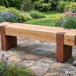 May include: A rectangular wooden bench featuring a thick, light-toned seat supported by two sturdy, dark-stained vertical legs. The construction includes visible metal brackets connecting the seat to the legs. The text MADE IN USA is visible.