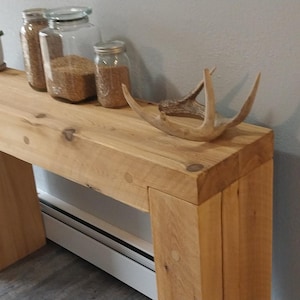 May include: A light-colored wooden console table with a rectangular top and two thick, square legs. The table is decorated with glass jars, a small potted plant, and a decorative antler. The table is approximately 36 inches long.