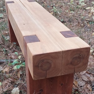 May include: A handcrafted wooden bench with a light brown seat and dark brown accents. The bench is made of thick, solid wood and features a simple, sturdy design. The bench is suitable for outdoor use.