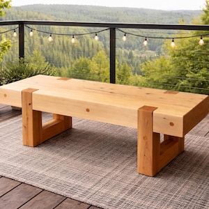 May include: A light-colored wooden bench with a rectangular top and four sturdy legs. The bench is made of natural wood and has a simple, modern design. It is placed on a gray rug on a wooden deck, with a scenic outdoor background.