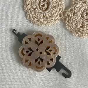 May include: A beige plastic yarn holder with a decorative floral design. The holder has a metal hook on each side.