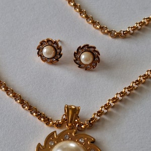 May include: A gold necklace with a pendant featuring a pearl surrounded by a swirl design and small clear stones. The necklace comes with a pair of matching earrings with pearls and clear stones.
