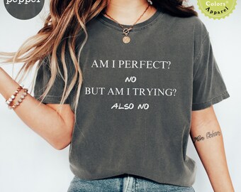 以下が含まれることがあります： 前面に「AM I PERFECT? NO BUT AM I TRYING? ALSO NO」という文字がプリントされた濃いグレーのTシャツ。長い茶色の髪をした人が着用しており、青いジーンズと合わせています。