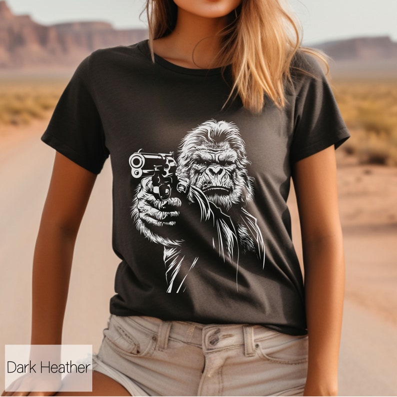 以下が含まれることがあります： 暗いヘザーグレーのTシャツに、銃を持ったゴリラの白いグラフィックがプリントされています。