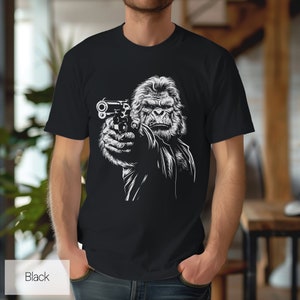以下が含まれることがあります： 銃を持ったゴリラの白いグラフィックがプリントされた黒いTシャツ