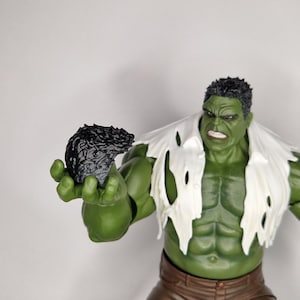 Könnte beinhalten: Eine grünhäutige Actionfigur mit kurzem schwarzem Haar und muskulösem Körperbau, bekleidet mit einem zerrissenen weißen Hemd und einer braunen Hose. Die Figur hat einen wütenden Gesichtsausdruck und hält ein kleines, texturiertes schwarzes Objekt.