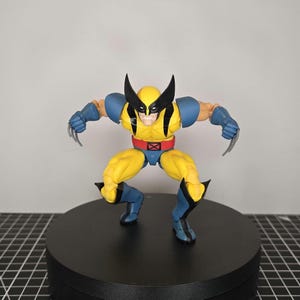 Peut inclure: Figurine d'action jaune et bleue d'un super-héros avec des détails noirs, des griffes et un masque. La figurine est posée de manière dynamique, les bras tendus et les jambes pliées. La figurine est sur un socle circulaire noir.