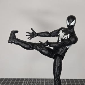 May include: A black action figure of Spider-Man in a dynamic pose, with one leg extended and arms outstretched. The figure has a white spider emblem on the chest and white eye details. The figure is on a gray surface.