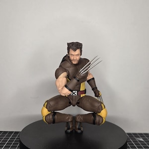 Może przedstawiać: Figurka Wolverine w pozycji kucznej, z wysuniętymi metalowymi pazurami. Figurka ubrana jest w brązowo-żółty kostium z czarnym pasem. Figurka stoi na czarnej, okrągłej podstawie.