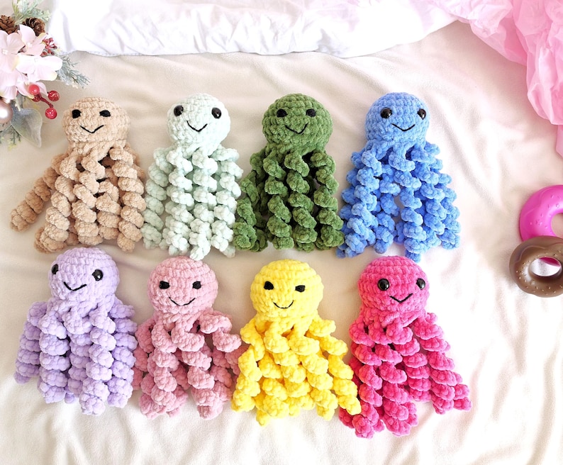 May include: Eight small, handmade crochet octopus plush toys arranged in two rows. Each octopus features a round head, black safety eyes, a stitched smile, and eight curly, spiraled tentacles made from soft, plush yarn in various solid colors.