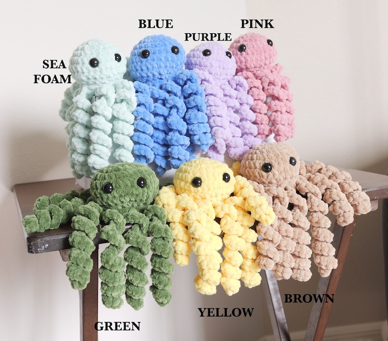 May include: A collection of seven crocheted octopus plush toys with black plastic eyes. The colors are labeled as SEA FOAM, BLUE, PURPLE, PINK, GREEN, YELLOW, and BROWN. Each octopus features a round head and eight curly, textured tentacles.