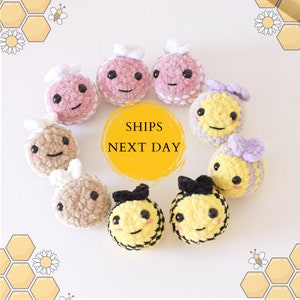 Peut inclure: Une collection de peluches abeilles et fleurs crochetées à la main. Les peluches sont faites dans différentes couleurs, dont le jaune, le rose, le violet et le marron. Les peluches ont des visages mignons avec des yeux et des sourires brodés. Les peluches sont disposées en cercle sur un fond blanc. Le texte "SHIPS NEXT DAY" est écrit en jaune sur un cercle jaune au centre de l'image.