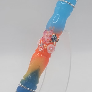 May include: A UV reactive pen with a blue and orange gradient design. The pen is decorated with silver-coloured bands and sea-themed embellishments, including a starfish and a turtle. The text "MM803 Reef Sunset Medium/Slim UV Reactive" is visible.