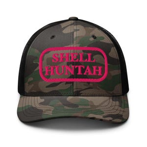 May include: Camouflage trucker hat with a black mesh back and a curved brim. The front panel features the text "SHELL HUNTAH" in bright pink, enclosed in a pink rectangular border. The hat is designed for outdoor activities.